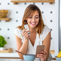 A smiling woman in a white shirt holds a Biscuits Fidget Slider Cartoon Haptic Toy while looking at her smartphone at the kitchen counter, possibly checking sensory toy certifications in China.