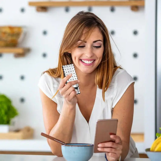 A smiling woman in a white shirt holds a Biscuits Fidget Slider Cartoon Haptic Toy while looking at her smartphone at the kitchen counter, possibly checking sensory toy certifications in China.