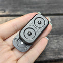 A hand holds the Owl Push Slider Fidget Slider Toy, a small black and gray metal EDC haptic fidget with two circular indents and a central button, against a wooden background.