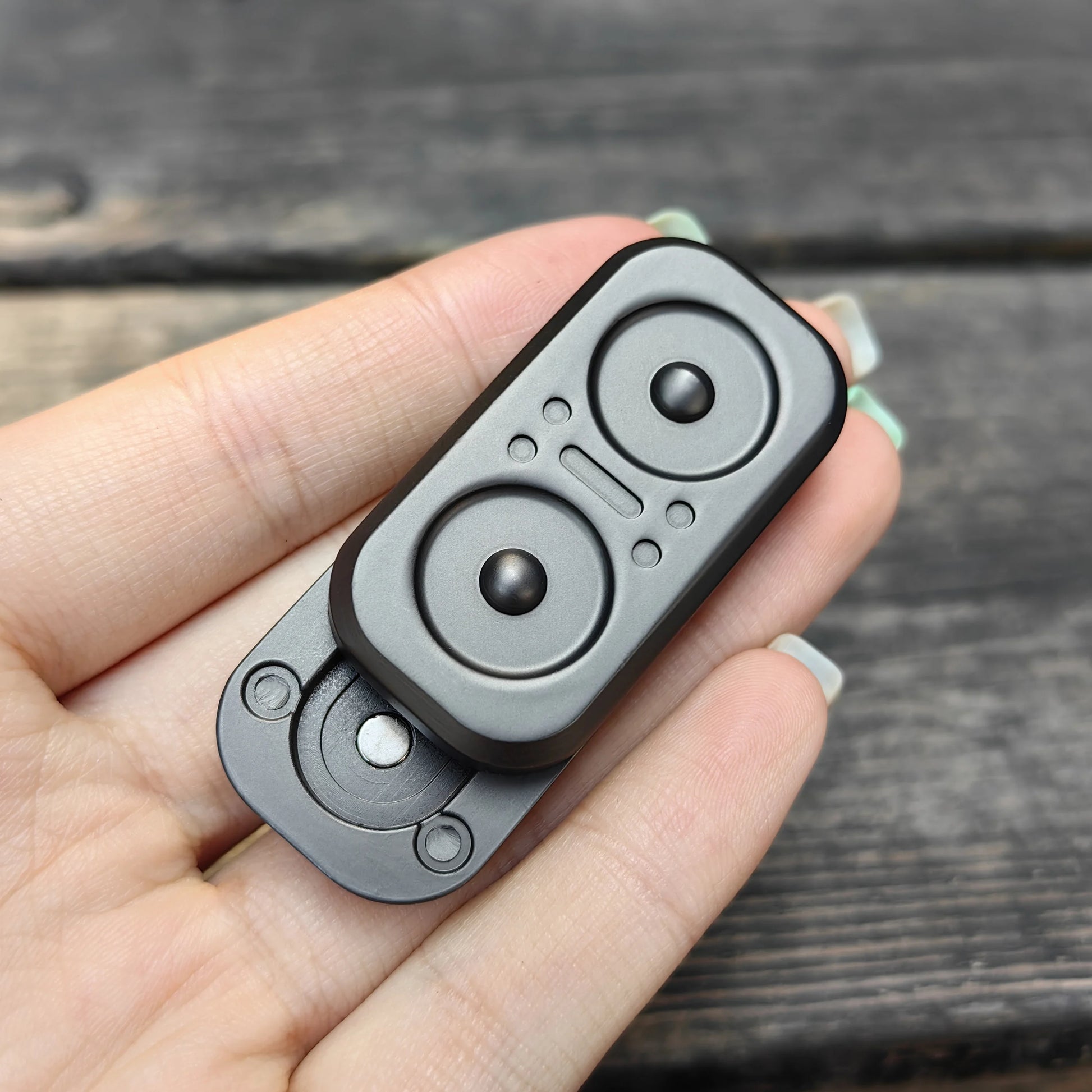 A hand holds the Owl Push Slider Fidget Slider Toy, a small black and gray metal EDC haptic fidget with two circular indents and a central button, against a wooden background.
