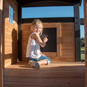 A young girl smiles as she draws a heart on a chalkboard inside the Playhouse Backyard Discovery Canyon Creek Cedar Swing Set, with sunlight streaming through the window and creating a warm, cheerful atmosphere.