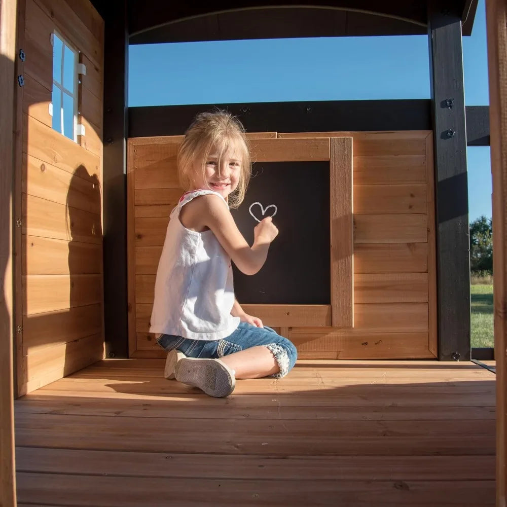 A young girl smiles as she draws a heart on a chalkboard inside the Playhouse Backyard Discovery Canyon Creek Cedar Swing Set, with sunlight streaming through the window and creating a warm, cheerful atmosphere.