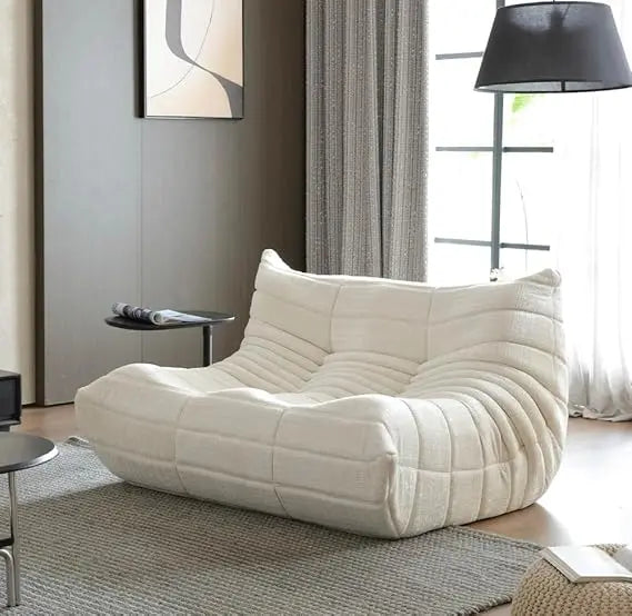 A modern living room featuring the Fireside Loveseat Sofa Couch in Chenille Beige, accented by a round side table, gray rug, tall curtains, floor lamp, and abstract wall art, all illuminated by natural light from large windows.
