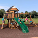 Children play on the Playhouse Backyard Discovery Canyon Creek Cedar Swing Set, which features a green slide, swings, climbing wall, and playhouse. Some swing or climb while others sit at a table beneath this outdoor playground set. Trees and grass surround them.