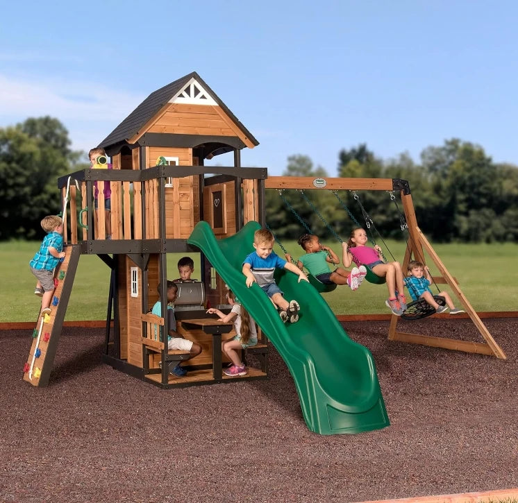 Children play on the Playhouse Backyard Discovery Canyon Creek Cedar Swing Set, which features a green slide, swings, climbing wall, and playhouse. Some swing or climb while others sit at a table beneath this outdoor playground set. Trees and grass surround them.
