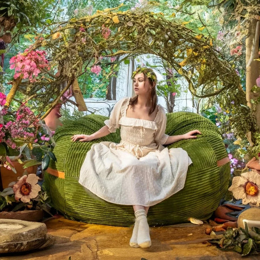 A young woman in a cream dress and flower crown sits on the Fuf XXL Foam Filled Bean Bag Chair, Moss Green, 6 Feet, beneath a floral arch and surrounded by oversized flowers in a whimsical fairy-tale garden.
