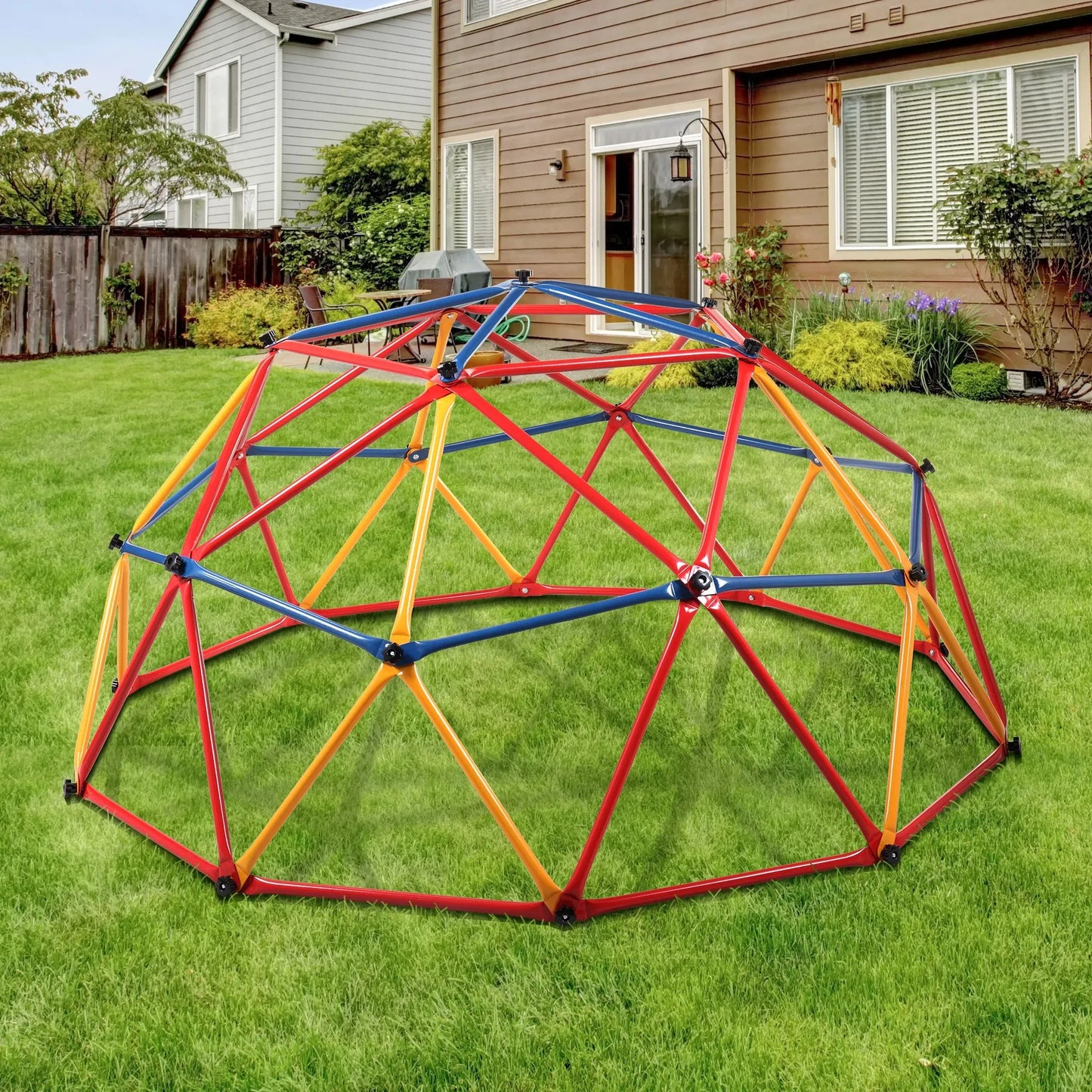 A Kids Outdoor Climbing Dome featuring red, yellow, and blue metal bars is installed on a grassy backyard near a brown house with white-framed windows and a wooden fence, providing an exciting play area for children.