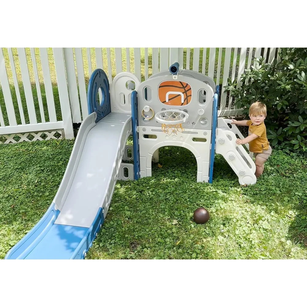 A toddler stands beside the 8 In 1 Toddler Slide Set With Basketball Hoop For Kids, which includes a slide, climbing wall, and basketball hoop on a grassy yard with a ball nearby and a white fence in the background.