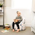 A young child with light hair sits barefoot on the Dolce Terazzo Lenox Milano Kids Bean Bag Chair for Comfort and Fun, holding an open book in a bright, cozy room with books, toys, soft carpeting, and a window.