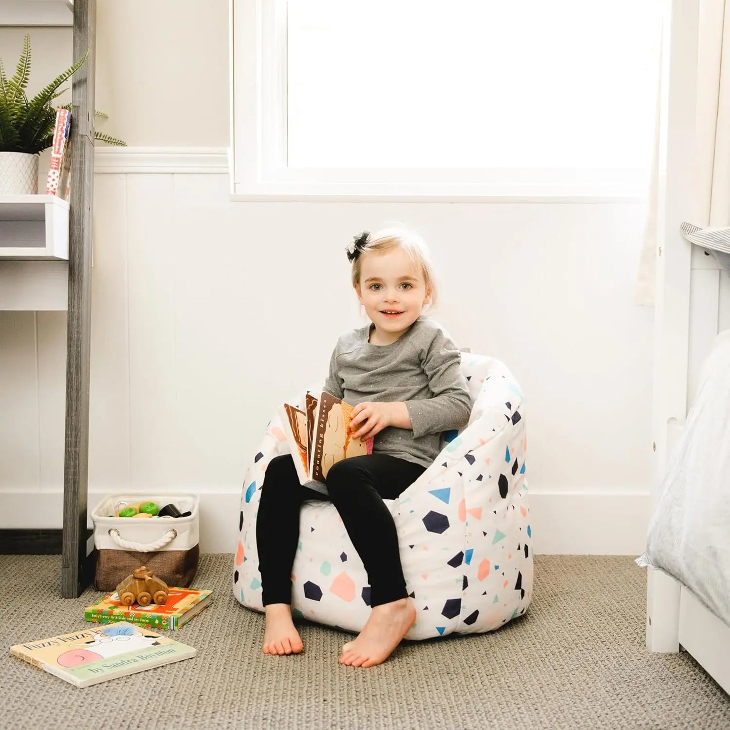 A young child with light hair sits barefoot on the Dolce Terazzo Lenox Milano Kids Bean Bag Chair for Comfort and Fun, holding an open book in a bright, cozy room with books, toys, soft carpeting, and a window.