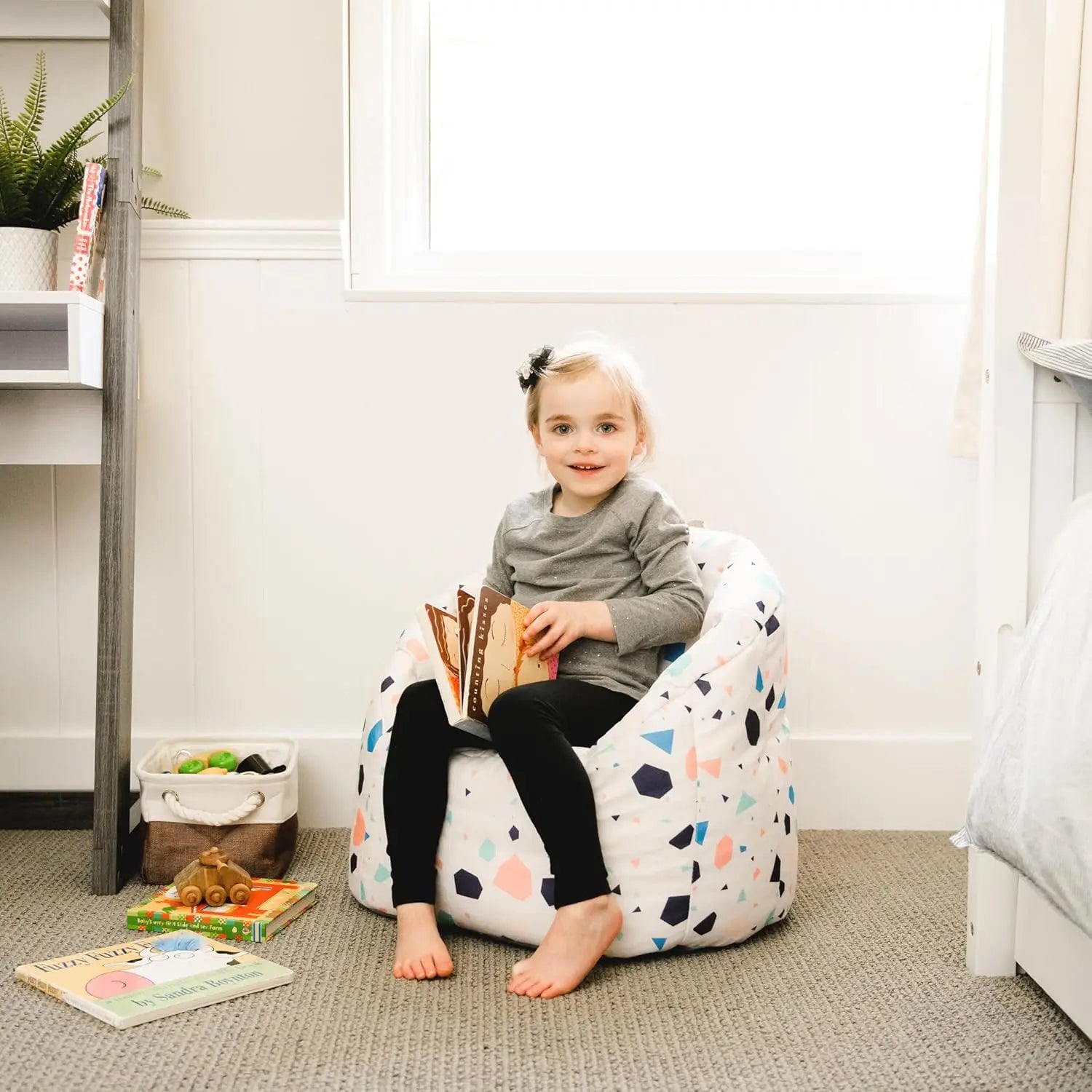 A young child with light hair sits barefoot on the Dolce Terazzo Lenox Milano Kids Bean Bag Chair for Comfort and Fun, holding an open book in a bright, cozy room with books, toys, soft carpeting, and a window.