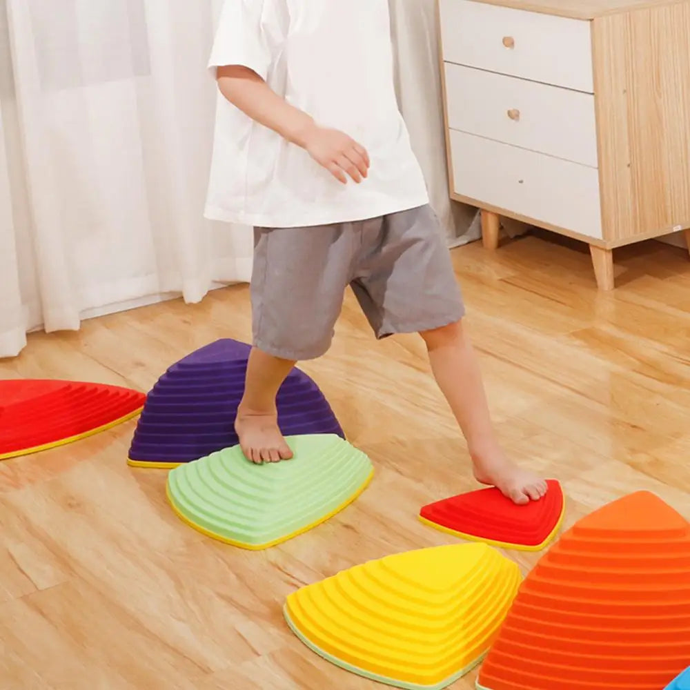 A child in a white shirt and gray shorts skillfully balances on Kids Balance River Stones, 5 non-slip sensory toys shaped like triangular domes, creating a vibrant obstacle course on a wooden floor with a light-colored dresser and white curtains in the background.
