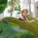 A woman in a floral crown and white dress lounges on a Fuf XXL Foam Filled Bean Bag Chair, Moss Green, 6 Feet, surrounded by lush indoor plants in a bright room with removable cover for easy care.