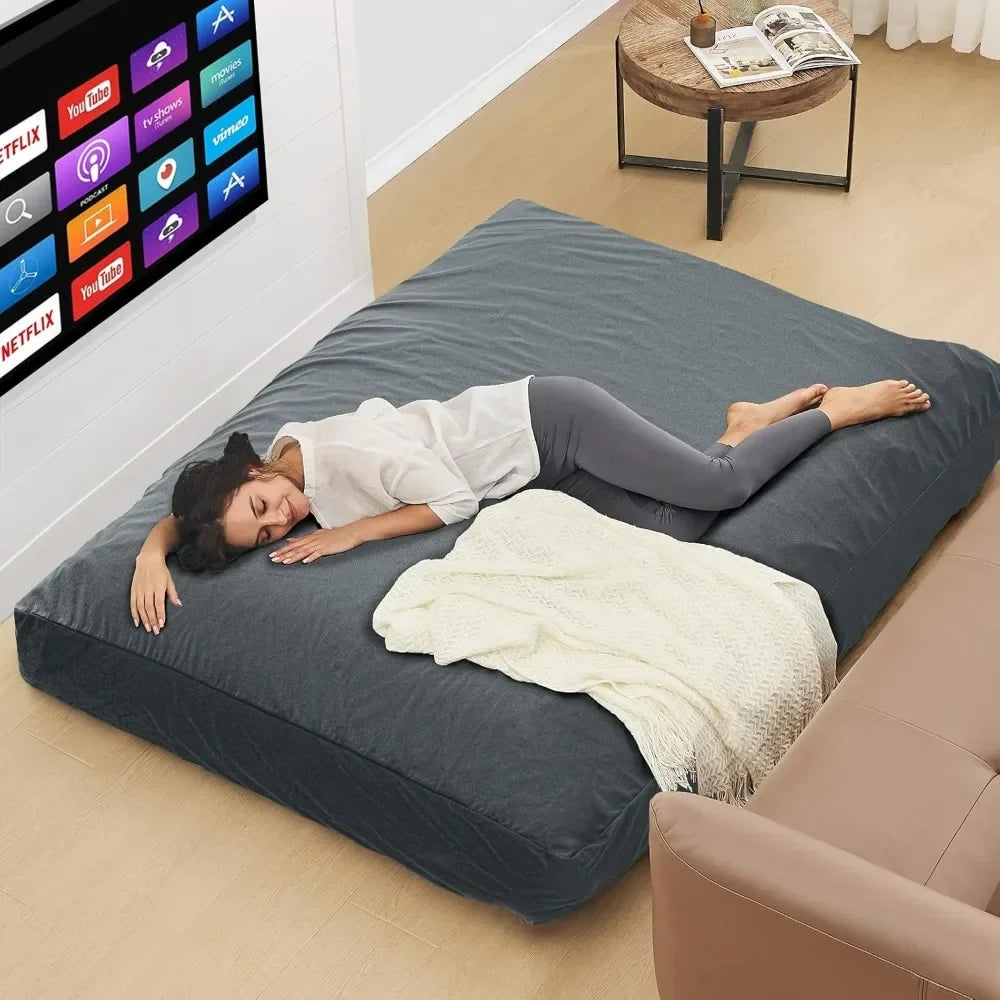 A woman naps on a large, dark gray Bean Bag Chairs Convertible Bean Bag Bed with a microfiber cover in a modern living room. A cream blanket lies nearby, and a TV with streaming apps is wall-mounted above a coffee table.