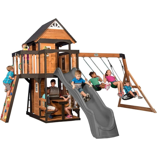 Kids have fun on the Playhouse Backyard Discovery Canyon Creek Cedar Swing Set, using the slide, swings, and climbing wall. Some relax at the picnic table under the playhouse while others explore or play on the balcony above.