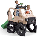 Two young children play on the Truck Climber Kids Outdoor Playset with Slide and Wall, shaped like a large beige toy truck with black wheels. One child climbs through the top holding a green toy; the other smiles inside. A green slide and climbing wall are attached.