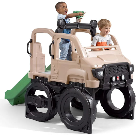 Two young children play on the Truck Climber Kids Outdoor Playset with Slide and Wall, shaped like a large beige toy truck with black wheels. One child climbs through the top holding a green toy; the other smiles inside. A green slide and climbing wall are attached.