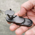 A hand holds the Target Push Slider Fidget Slider Toy, a small, sleek black metal EDC haptic slider for adult stress relief, featuring a circular pad and raised stand, set against a blurred gray background.