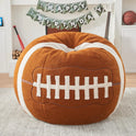The Sports Football Bean Bag Chair for Adults and Kids in Football color, crafted with ultra-soft sherpa plush, sits in a sports-themed living room decorated with a Happy Birthday banner, guitar, bat, basketball, and stacked firewood.