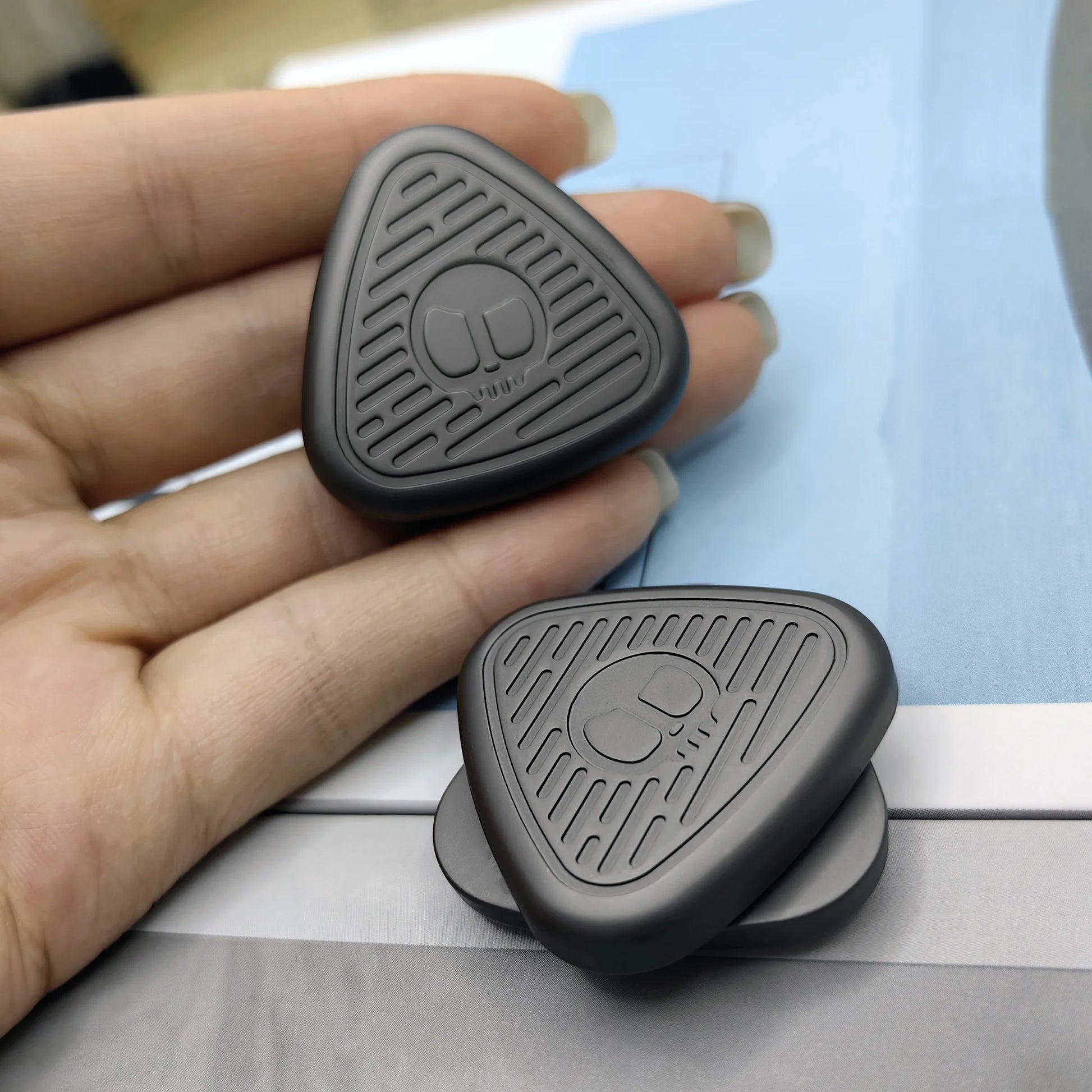A hand holds the gray EDC Fingertip Toys Fidget Slider, featuring textured grooves and a logo; another slider lies on a blue and white surface. Both magnetic metal fidget spinners are crafted in Mainland China.