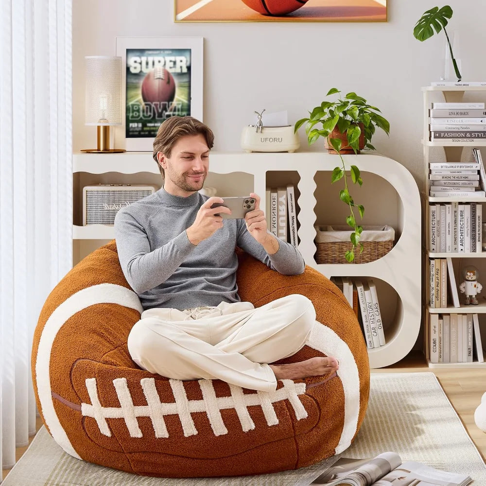 A man relaxes on a Sports Football Bean Bag Chair for Adults and Kids, smiling at his phone. The modern room with bookshelves, plants, and a Super Bowl poster creates the ideal space for sports fans.