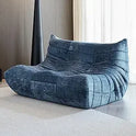 A modern Fireside Loveseat Sofa Couch in Chenille Blue with an ergonomic, curved, quilted design sits on a light rug in a contemporary room with neutral walls and sheer white curtains.