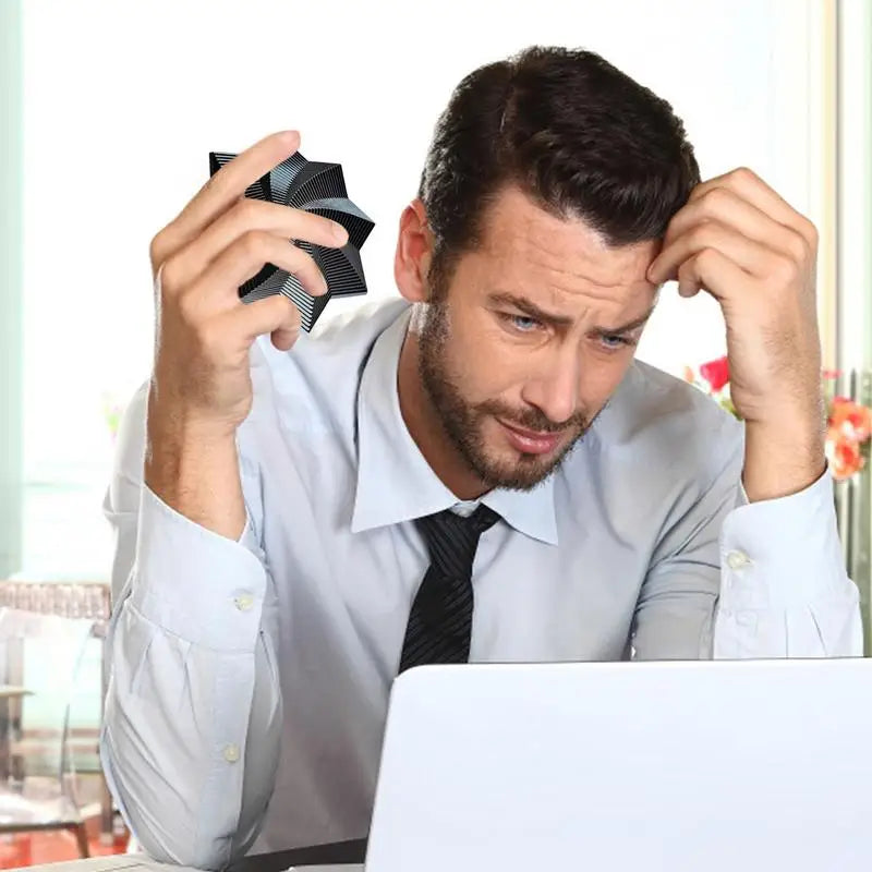 A man in a shirt and tie sits at a desk, looking stressed while holding his head with one hand and a Fidget 3D Spinner Stress Relief Toy in the other, in front of an open laptop.