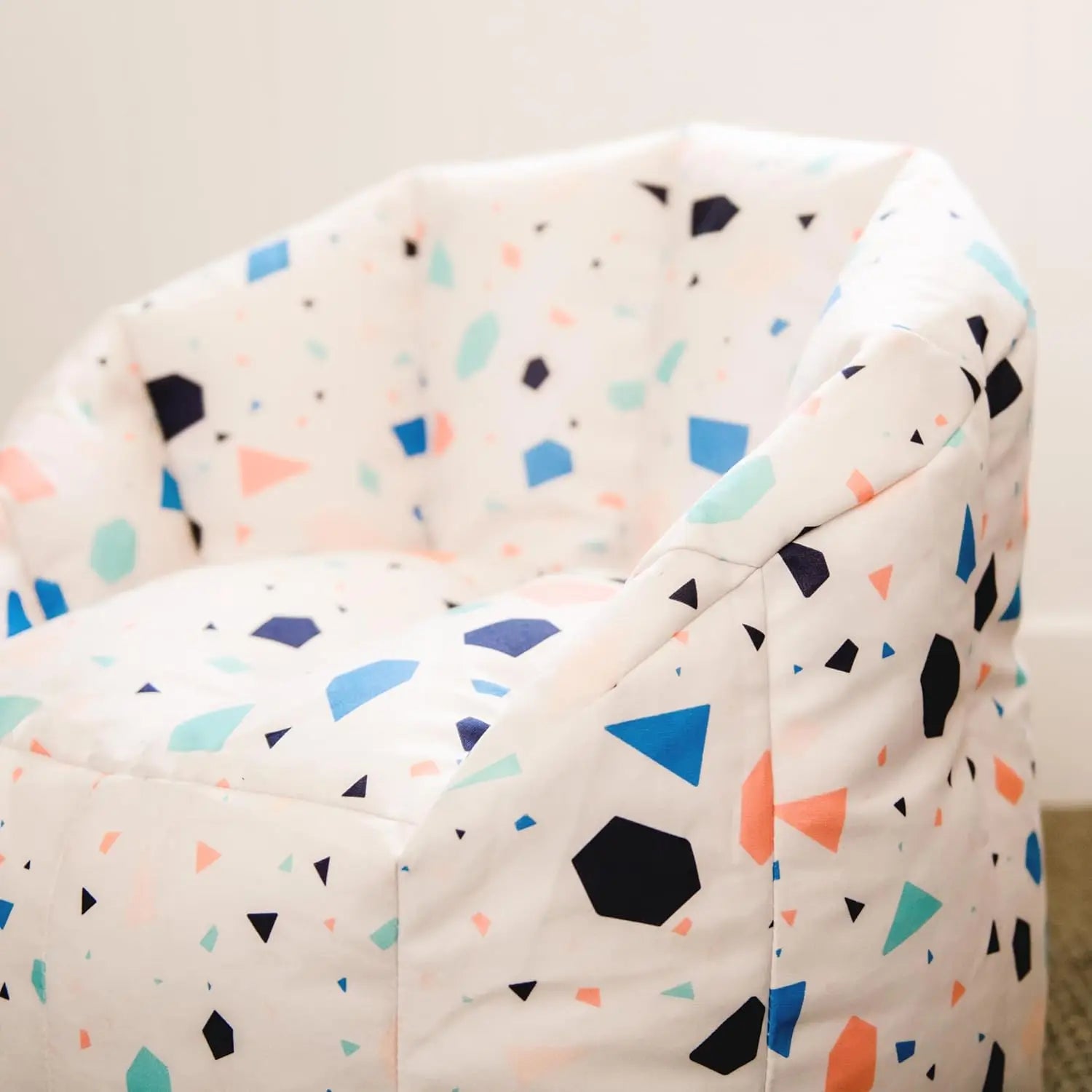 The Milano Kids Bean Bag Chair for Comfort and Fun features white, kid-friendly fabric with a blue, teal, coral, and black geometric pattern. Ideal for kids’ rooms, it’s shown indoors against a white wall.