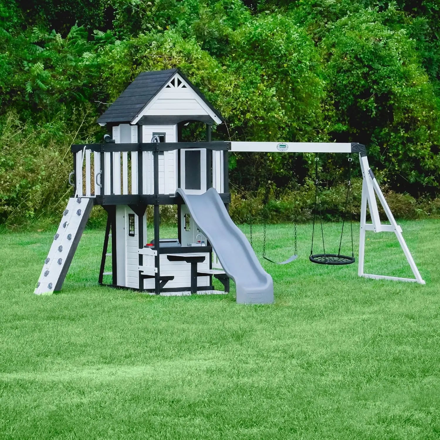 A white Playhouse Backyard Discovery Canyon Creek Cedar Swing Set on grass, featuring a playhouse, gray slide, climbing wall, two swings, picnic table underneath, and surrounded by trees—ideal for outdoor fun.