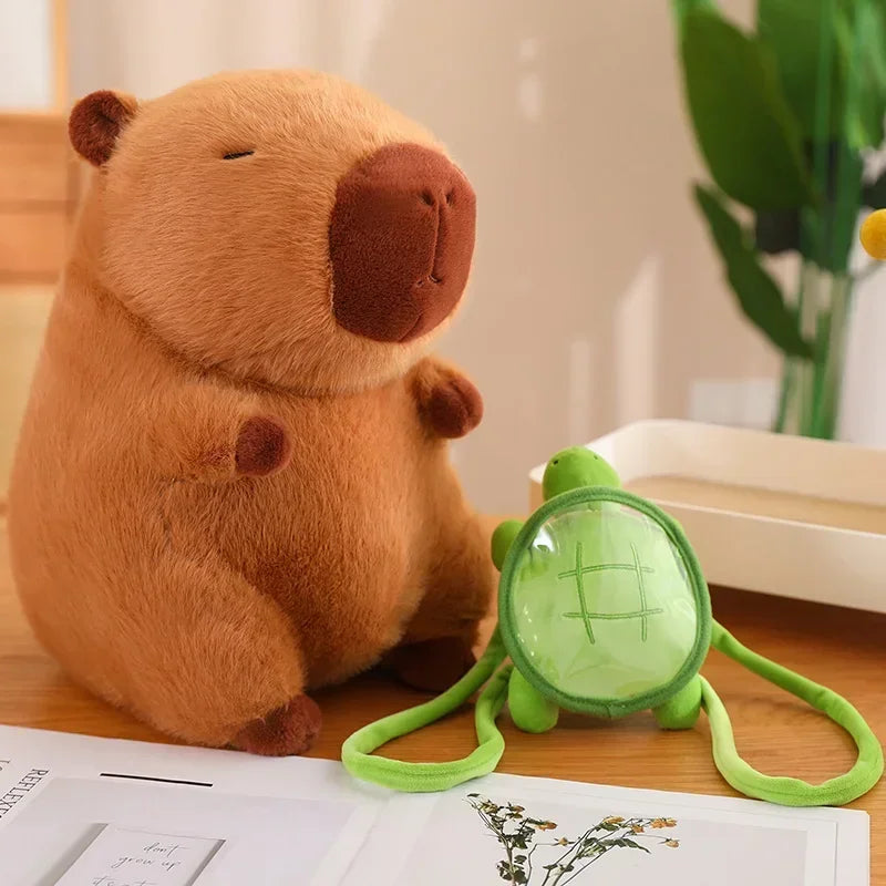 A Cute Turtle Backpack Capibara Strawberry Hat Capybara Plush Toy sits on a table beside a small green turtle stuffed animal with a transparent shell, with papers and a plant in the background.