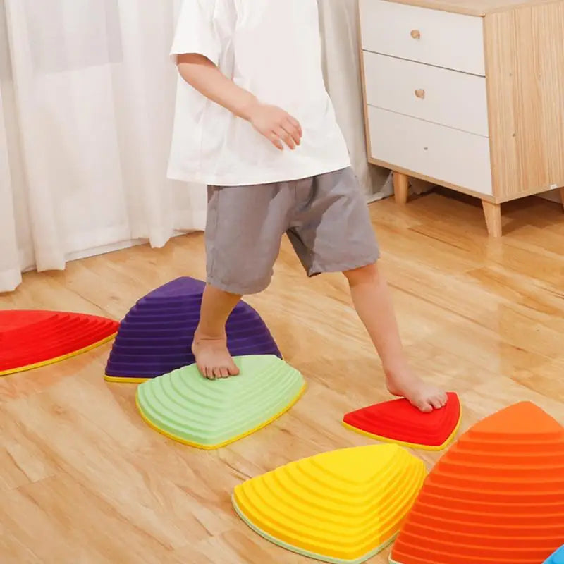 A child in a white shirt and gray shorts balances on colorful Kids Balance River Stones—five non-slip sensory toys in red, yellow, orange, green, and purple—arranged on a wooden floor. A dresser is visible in the background.