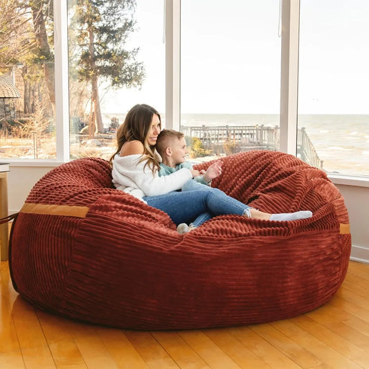 A woman and a child relax on a Fuf XXL Foam Filled Bean Bag Chair, Deep Terracotta 6 Feet, enjoying its cozy comfort in a sunlit room with large windows overlooking a scenic lake and trees.