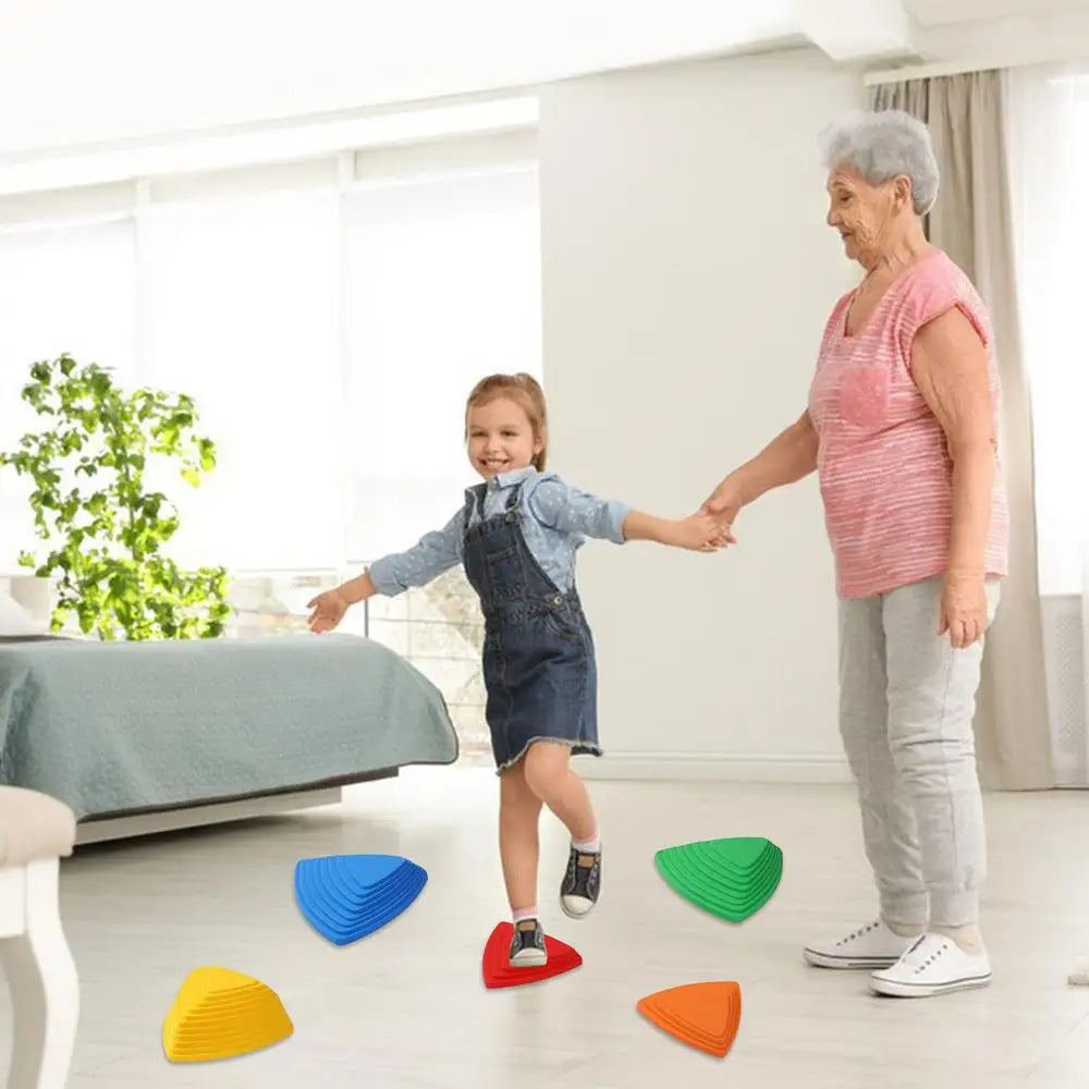A smiling child in blue dress and denim overalls plays on colorful Kids Balance River Stones with an older woman in a pink top and gray pants. They are indoors, crafting a fun obstacle course with a plant and bed in the background.