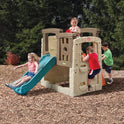 Four young children play outdoors on the Woodland Climber II Kids Playset for Ages 2 to 6 Years. One girl slides down the toddler slide, while others climb and explore, surrounded by trees and wood chips.