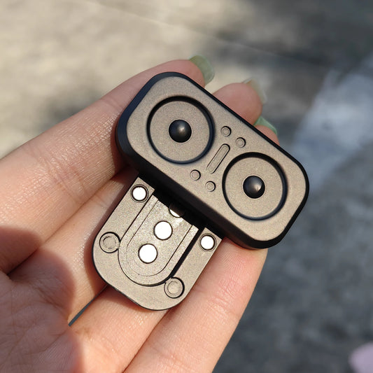 A hand holds the Owl Push Slider Fidget Toy, a compact metallic device with two black circles like eyes, resembling an owl’s face. Made from metal, this unique EDC haptic slider is perfect for finger spinning and stress relief. Blurred pavement in the background.