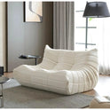 A modern living room showcases the cream-colored Fireside Loveseat Sofa Couch Modern Chenille Bean Bag Chair on a textured gray rug, beside a black floor lamp, with gray curtains, a window, and abstract wall art in the background.