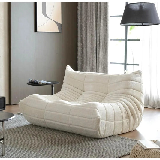 A modern living room showcases the cream-colored Fireside Loveseat Sofa Couch Modern Chenille Bean Bag Chair on a textured gray rug, beside a black floor lamp, with gray curtains, a window, and abstract wall art in the background.