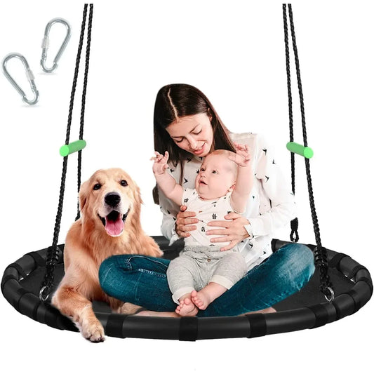 A woman sits on a 40-inch Tree Swing with PP Mat for Kids and Adults, Durable Black Outdoor Saucer Swing, holding a happy baby and smiling golden retriever. Two metal carabiners are visible in the top left corner.