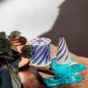 Two small Impossible Cone Sensory Fidgets with white rubber bases and swirling purple patterns—one cylindrical, one cone-shaped—rest on a wooden surface near a potted plant and decorative blue and amber glass pieces.