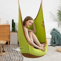 A woman relaxes in a Kids Pod Swing Seat Bag Sensory Swing Portable Hammock Chair with cushion indoors, sunlight streaming in. The cozy room features a wooden dresser, houseplants, a white sofa with green throw, and a rug.