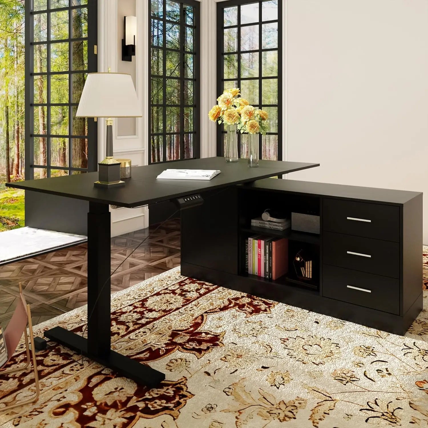 A modern home office with a black L Shaped Electric Standing Desk with Drawers and Shelves, an industrial-grade steel frame, table lamp, vase with flowers, books, and storage. Large windows overlook woods; an ornate rug covers the floor.