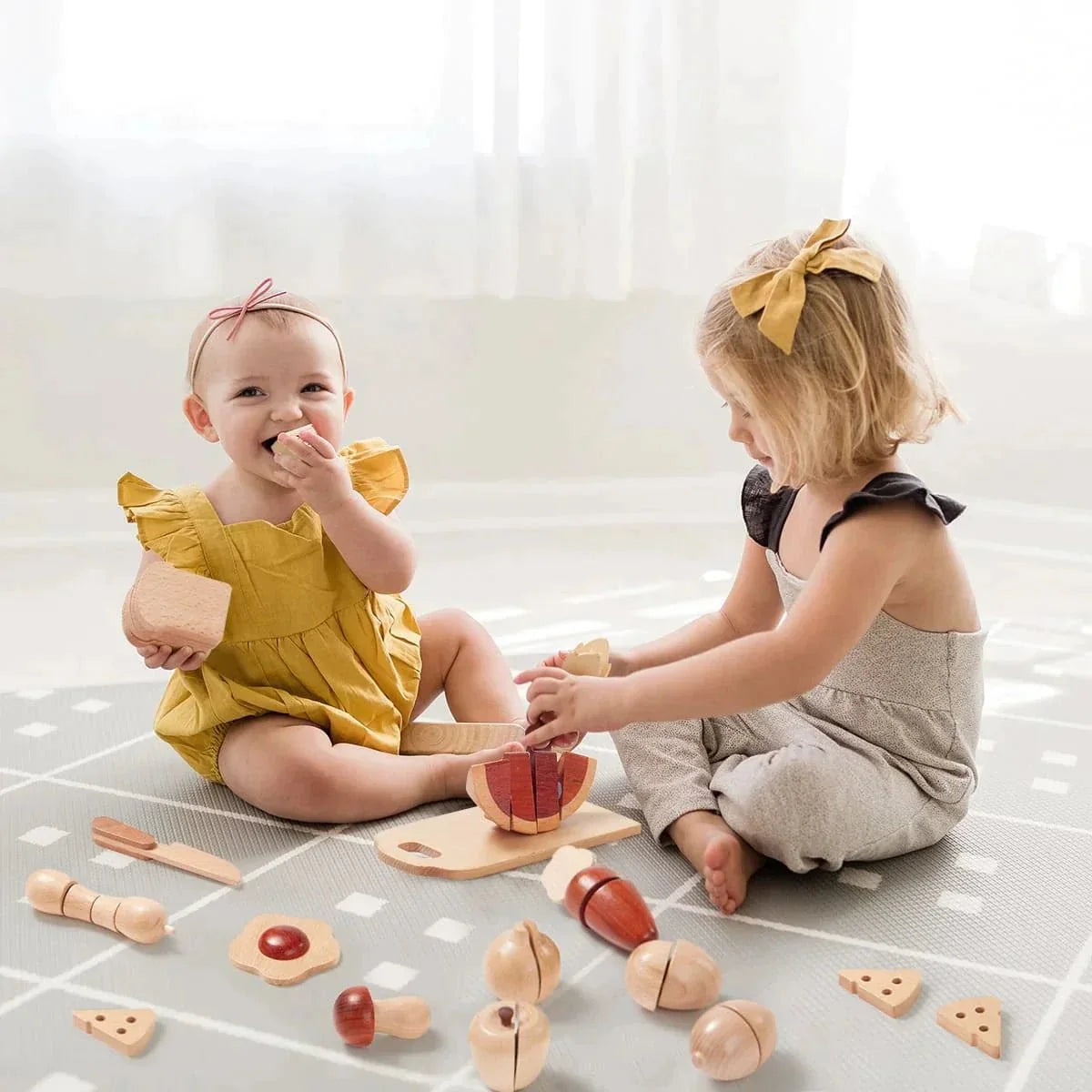 Two young children sit on a play mat playing with a Wooden Cut & Play Food Set with Knife and Board, which includes wooden fruits and veggies, a cutting board, and a toy knife.