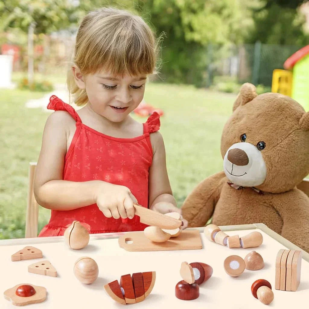 A young girl sits next to a teddy bear and uses a knife from the Wooden Cut & Play Food Set with Knife and Board to slice wooden fruits and vegetables on a cutting board.