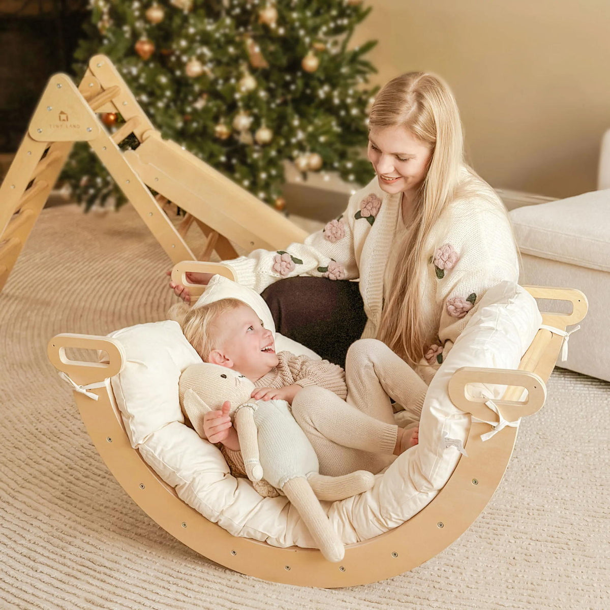 A woman sits on the floor watching a young child inside a curved wooden rocker lined with a Thick Padded Play Cushion (Arch Not Included).