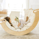 A child reads a book while sitting in a curved wooden rocking cradle lined with a cream-colored Thick Padded Play Cushion.