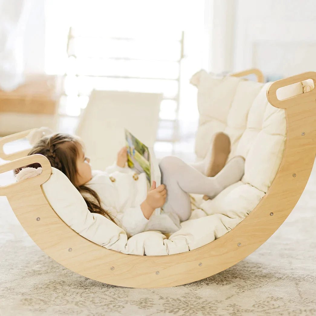 A child reads a book while sitting in a curved wooden rocking cradle lined with a cream-colored Thick Padded Play Cushion.