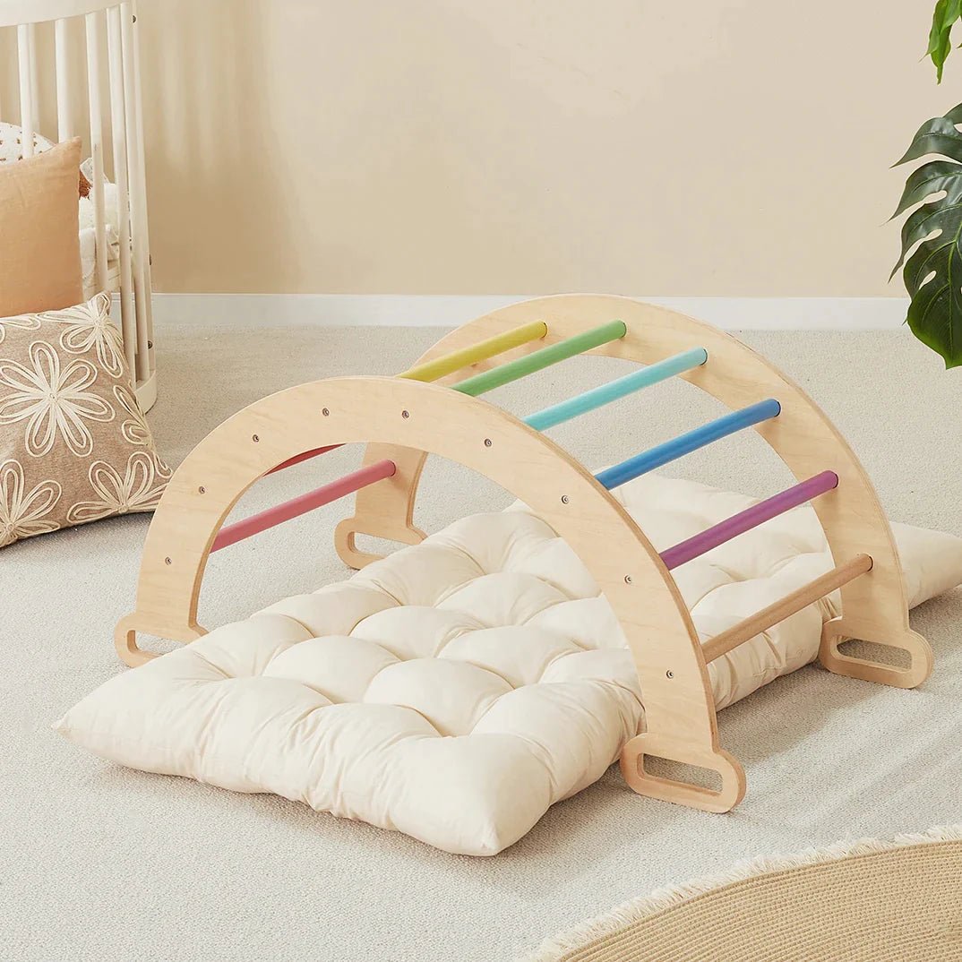 A wooden arched climbing frame with colorful rungs positioned over a Thick Padded Play Cushion, which is a tufted, rectangular off-white mat.