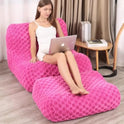 A woman in a white dress sits on the Widen-Pink Bean Bag Sofa with matching footstool, using a laptop. Indoors, she’s near books on a side table, with beige curtains in the background.
