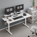 A 63-inch white Standing Desk w/ 2 Adjustable Monitor Shelves Electric Stand Up Desk features dual monitors showing “10:00,” keyboard, mouse, laptop, camera, plants, and office supplies. White drawers labeled 32 and a chair sit on light gray carpet nearby.