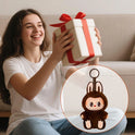 A smiling girl sits on the floor holding a wrapped gift with a red ribbon. An inset shows a Cute Labubu The Monsters Box Toy Sweetheart vinyl doll keychain, shaped like an adorable animal in a brown bunny costume.